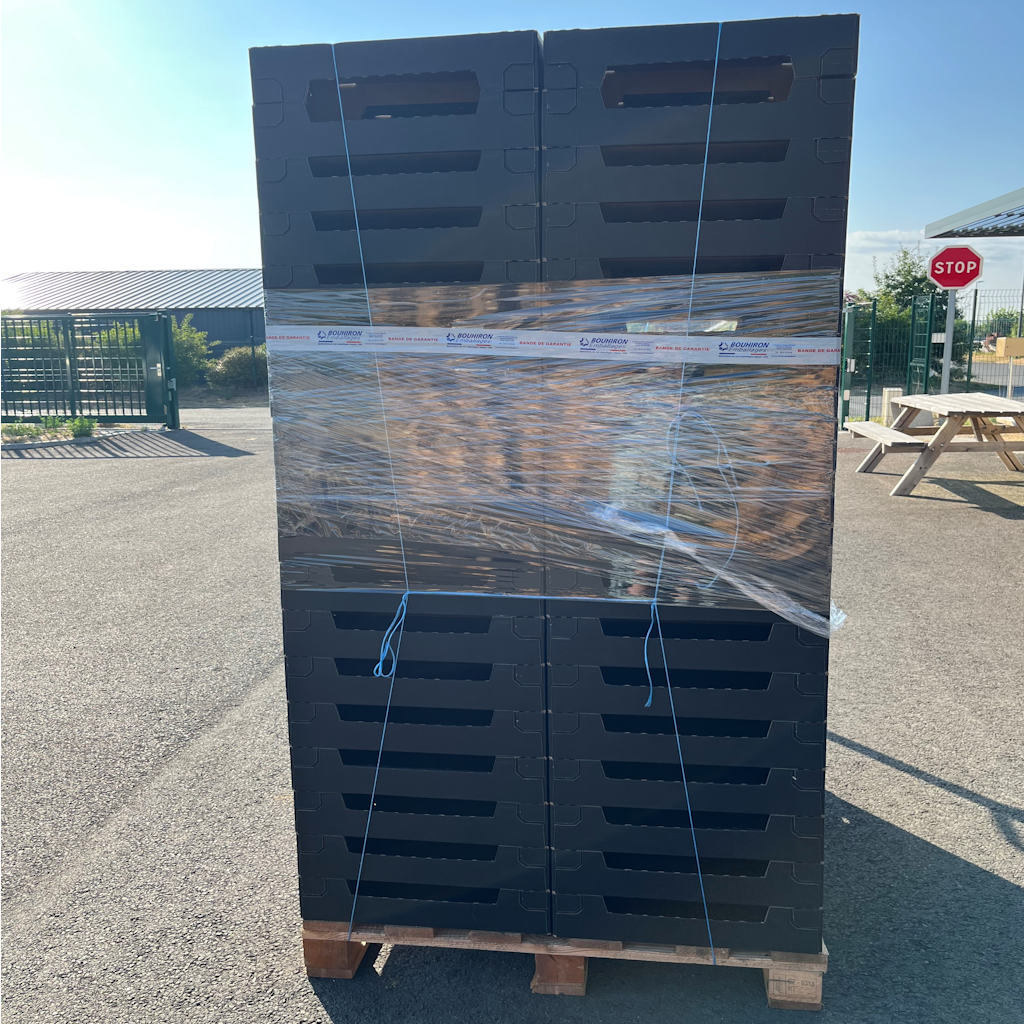 Palette de plateaux carton noirs empilés et filmés, entreposée dans une cour extérieure avec portail et panneau stop
