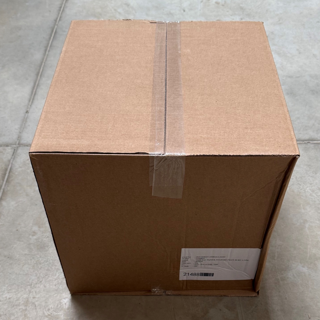 Carton d'expédition fermé contenant 250 sacs papier fenêtre filet écru 1,5kg, posé sur carrelage gris